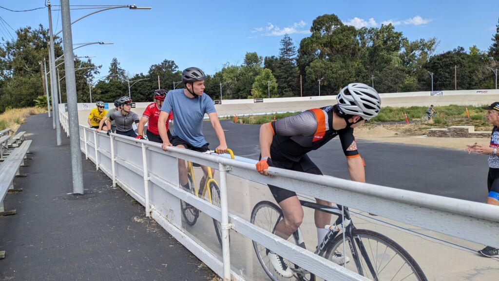 Several cyclists prepare to ride around a velodrome by holding the top rail and listening to their instructor.