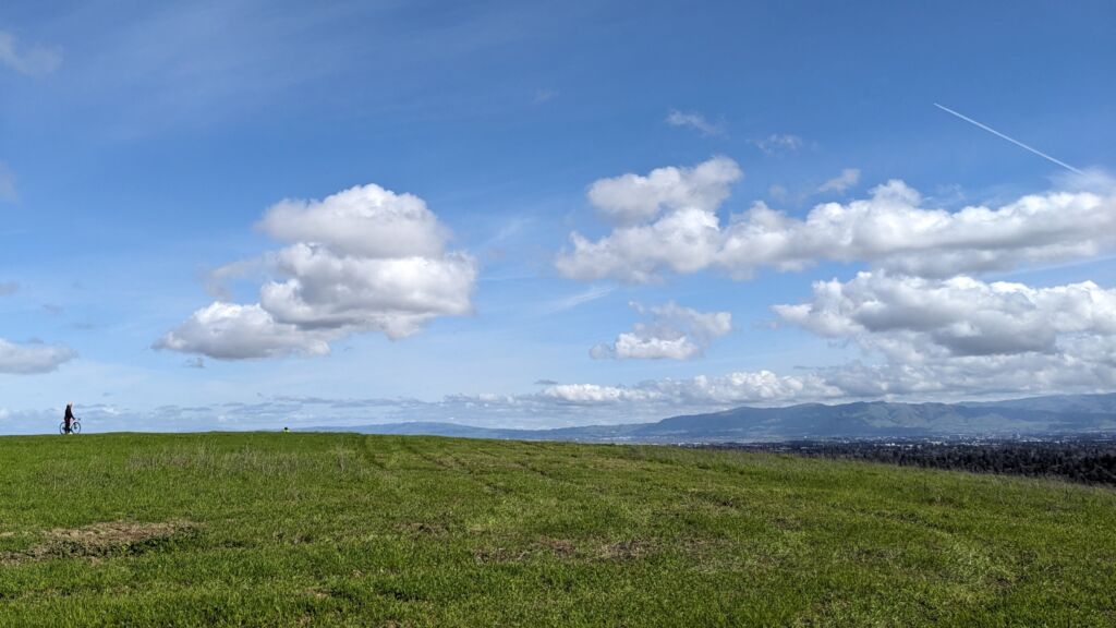 A lone cyclist on a grassy knoll overlooking a populated valley on a mostly sunny day.